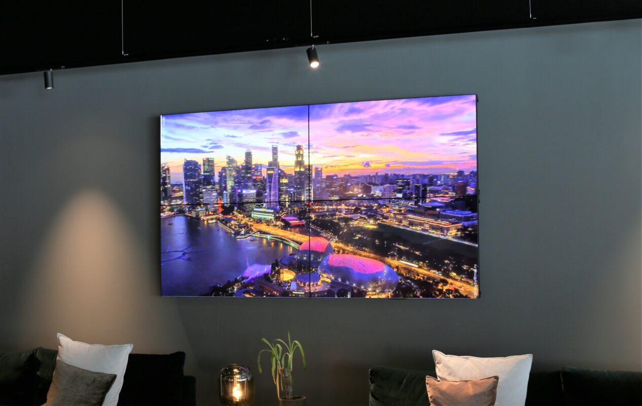 En stor platt-tv monterad på en grå vägg visar en stadsbild i solnedgång ovanför ett modernt vardagsrum, som visar upp smarta designlösningar med snygga kuddar och en liten växt.
