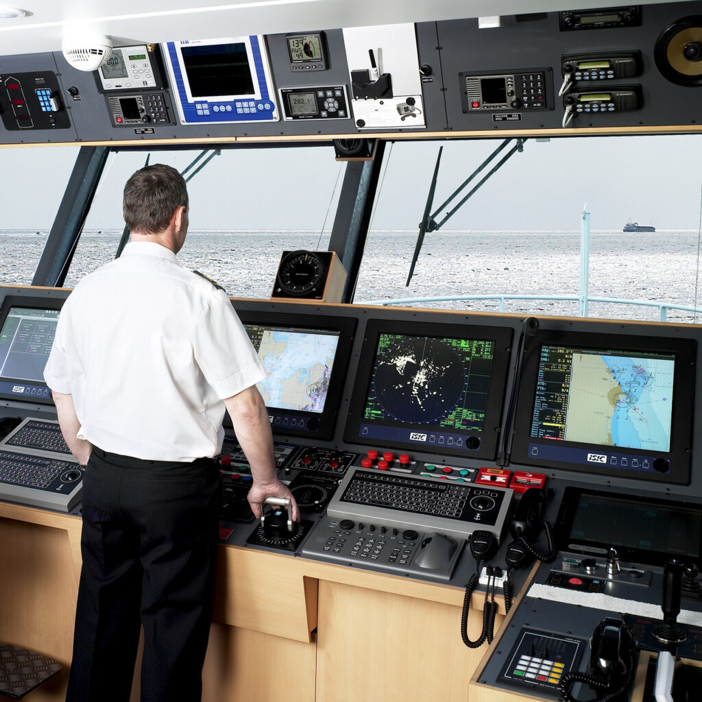 En man i vit uniform manövrerar en fartygsbrygga och står vid en kontrollpanel med flera skärmar och navigationsutrustning från Lagercrantz Group och tittar ut över havet.