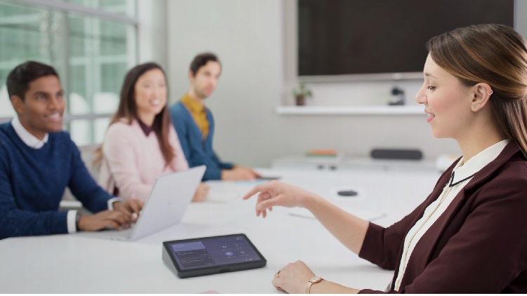 En kvinna i ett mötesrum gestikulerar mot en pekskärmsenhet med Microsoft Teams på bordet medan tre kollegor lyssnar, en av dem med en bärbar dator.