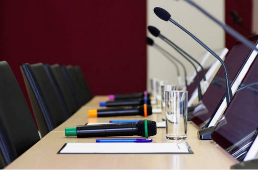 Ett konferensbord med tolkanläggningar, mikrofoner, anteckningsblock, pennor och vattenglas placerade framför stolar.