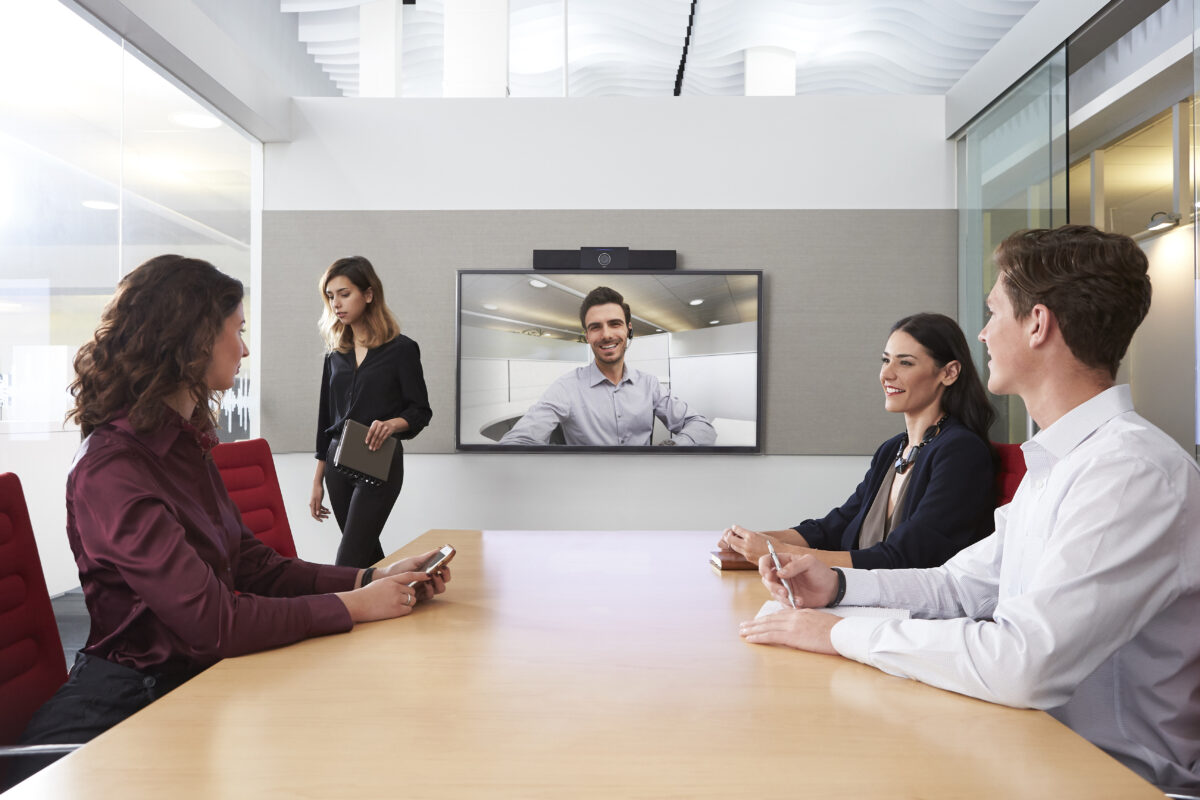 Fyra personer i ett konferensrum har en videokonferens med en man på en stor skärm; en person kommer in i rummet med en surfplatta.
