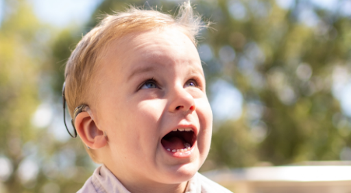 Ett litet barn som bär hörapparat tittar uppåt med öppen mun utomhus en solig dag, möjligen efter att ha upplevt Visual Reinforcement Audiometry (VRA) under en audiometri.