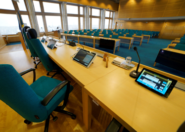 Ett modernt auditorium med rader av turkosa stolar, digitala surfplattor på träbord, mikrofoner och stora fönster som släpper in naturligt ljus.