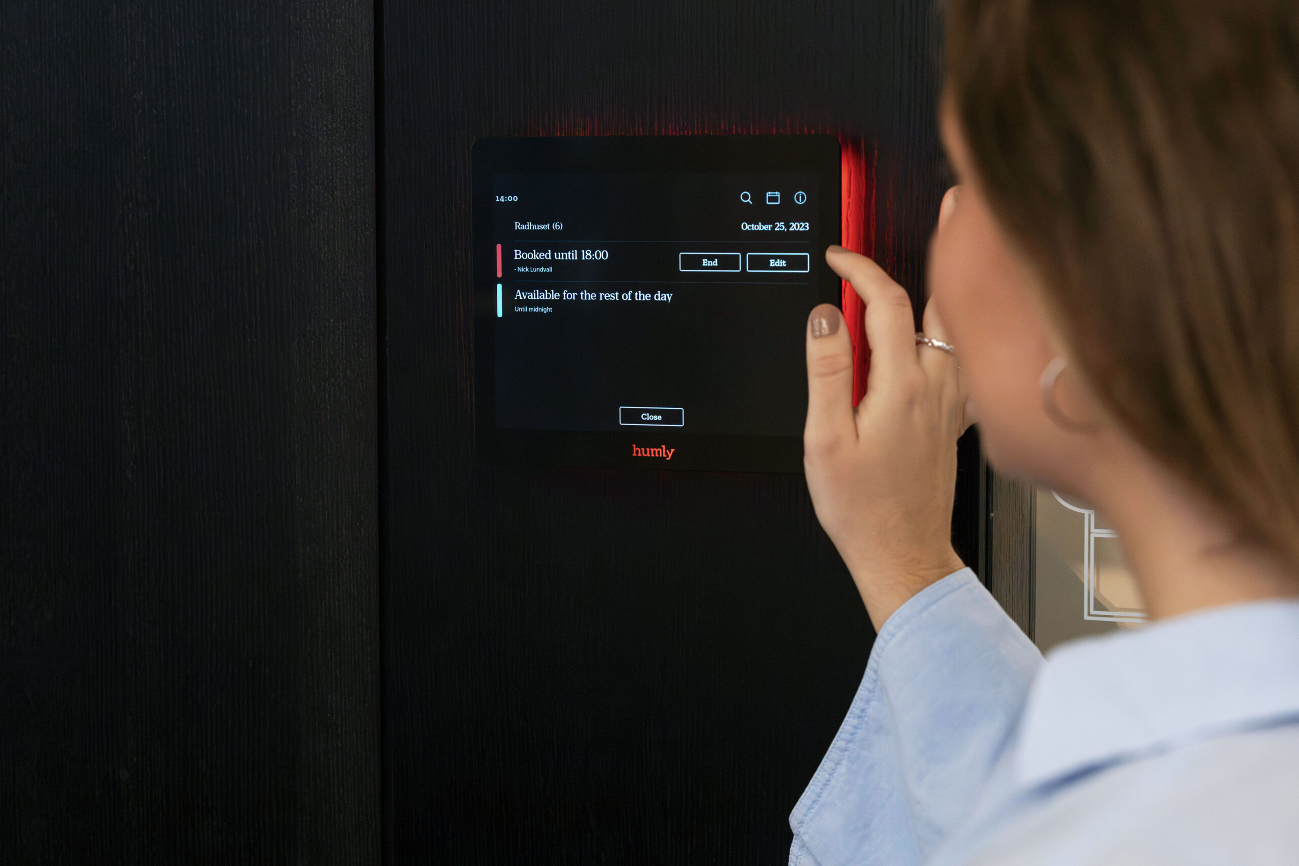En person interagerar med en väggmonterad Bokningsdisplayer, en digital schemaläggningspanel som visar mötesrummets tillgänglighet och bokningsalternativ.