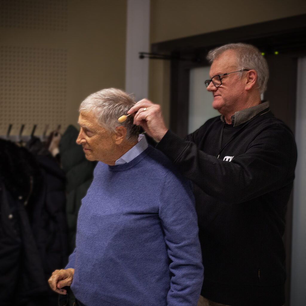 Tekniker justerar en liten klämmmikrofon på en äldre mans krage inför ett evenemang, stående bakom honom i en inomhusmiljö.