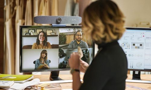 En person står vid ett skrivbord med en kopp i handen, vänd mot en datorskärm med tre personer i ett videosamtal som diskuterar Lösningar, medan ett designprogram är öppet på en annan skärm. En person står vid ett skrivbord med en kopp i handen, vänd mot en datorskärm med tre personer i ett videosamtal som diskuterar Lösningar, medan ett designprogram är öppet på en annan skärm.